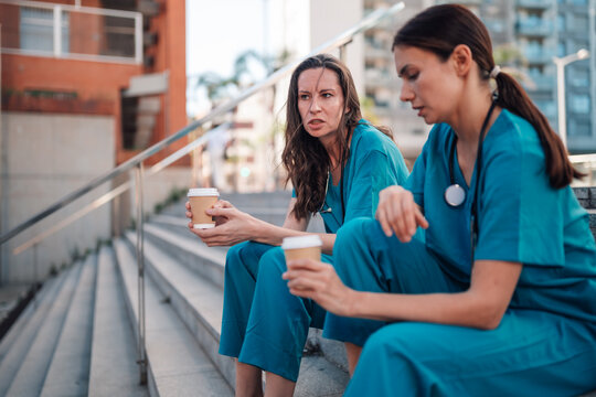 Tired female doctors in medical scrubs sitting on outdoor stairs, drinking coffee, and discussing work during a break