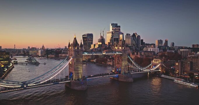 Famous Tower Bridge glowing with lights at golden colorful sunset. Reflecting in the River Thames with modern London cityscape and its financial district skyline in the background. Aerial drone shot