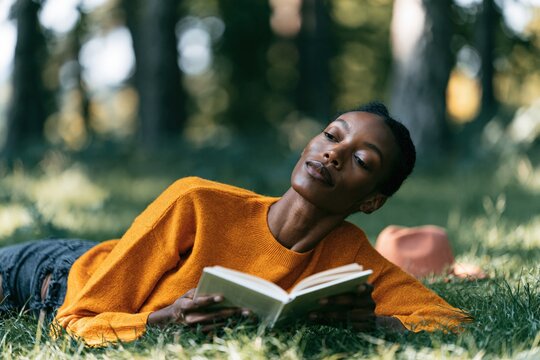 Woman reading book outdoors lying on grass in orange sweater for cozy lifestyle background and mindful relaxation banner