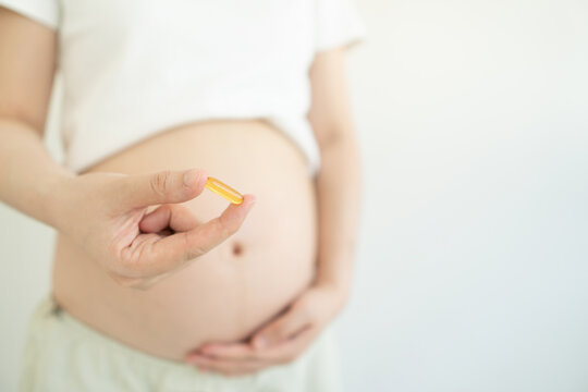 Pregnant woman holding dietary supplement capsule against white background. Omega 3 fatty acid DHA to support a healthy pregnancy and development of baby brain. Maternity prenatal care concept.