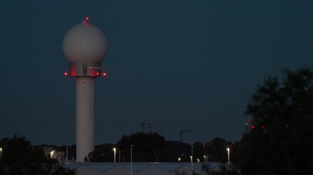 Spherical airport radar dome stands at night, red warning lights glowing at its base, surrounded by dark tree silhouettes and distant airport and city lights below
