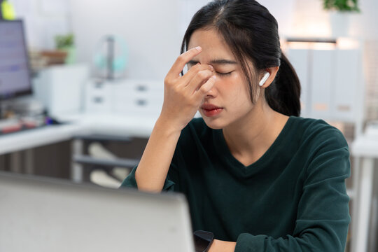 stressed young Asian businesswoman sits at her office desk with her hands on her forehead while looking at a laptop screen.image represents workplace stress, mental fatigue, burnout, deadline pressure