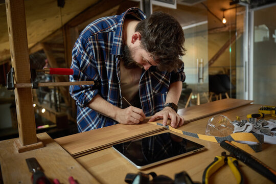 Focused bearded craftsman measuring wooden board beside tablet at garage workbench. Concept of digital woodworking guide, precision cutting preparation, DIY project tutorial.