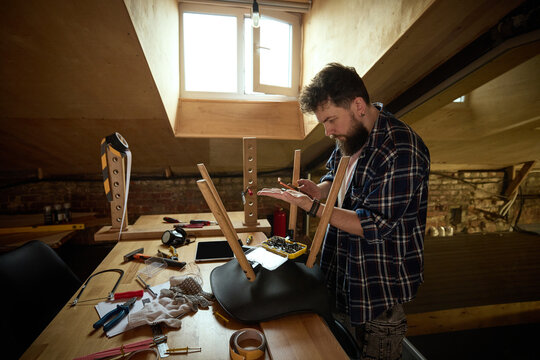 Bearded DIY worker following tablet instructions while assembling chair in garage. Concept of interactive assembly manual, furniture setup support, online maker education.