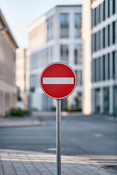 A red circular no entry traffic sign on a metal pole stands centered on a quiet city street, framed by modern office buildings and softly blurred pavement in clear daylight in a contemporary district
