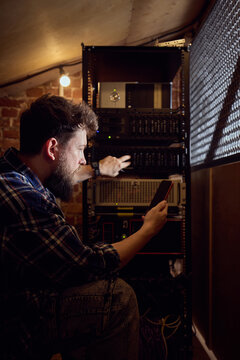 Male IT technician checking server rack with smartphone in dim technical room. Concept of data center maintenance, mobile network monitoring, IT support service.