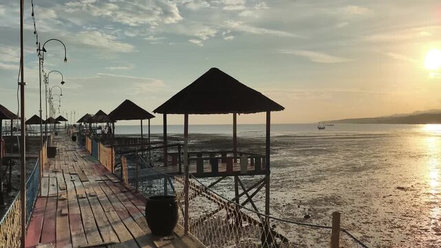 Wooden pier with thatched huts standing over mudflats at tropical coast during golden sunset. 4K Video