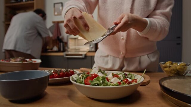 Close-up shot of unknown young woman grating cheese over fresh salad in kitchen while man cooking in background. Footage for healthy eating and meal kit delivery promotions