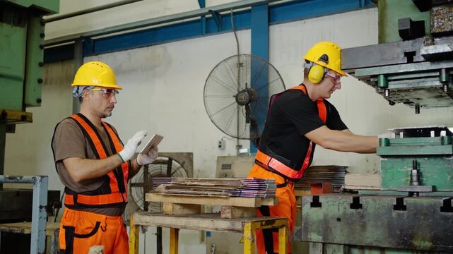 Two professional factory workers in PPE hardhats and vests operate a metal pressing machine and use a digital tablet for quality control inspection inside a heavy industrial manufacturing facility.