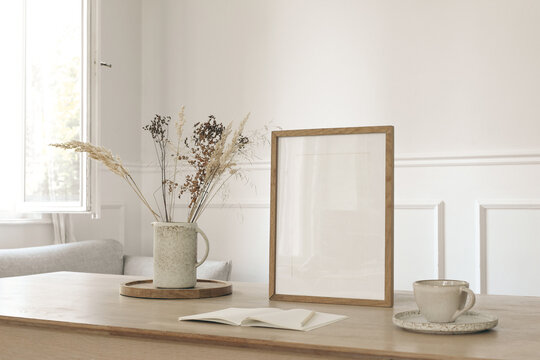Minimalist Home Interior with Blank Wooden Picture Frame Mockup. Ceramic Vase with Dried Flowers, Grass, Coffee Cup and Open Notebook on Wooden Table near Window. Blurred Living Room. Poster template