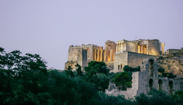 The Acropolis of Athens illuminated at dusk, with the Parthenon and surrounding ancient structures rising above trees and historic ruins.