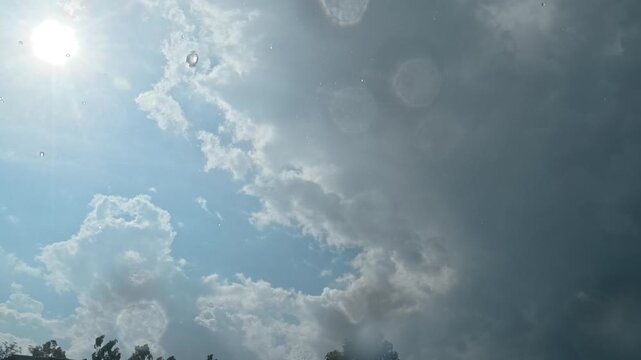 Raindrops fall on a waterproof camera as the sun shines through a cloudy sky, creating rainbow colors in some droplets.
