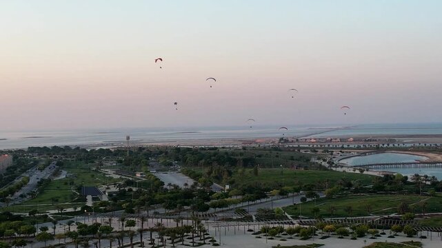 Wide aerial view of motorized paragliders flying over coastal park and sea during sunset
