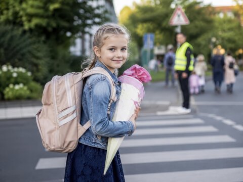 Sicherer Schulweg zur Einschulung in Deutschland: M&auml;dchen mit Schulranzen und Schult&uuml;te wartet am Zebrastreifen, w&auml;hrend ein Sch&uuml;lerlotse den Verkehr sichert.