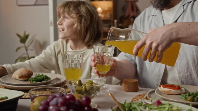 Caucasian man pouring orange juice from carafe into glass during family dinner next to child. Footage for promoting healthy family nutrition or organic beverages
