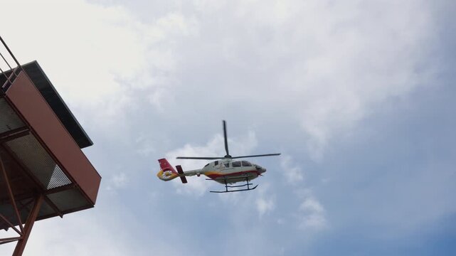 A red white and yellow helicopter flies overhead seen from a low angle beside the edge of a rust coloured steel observation tower, under a bright cloudy sky