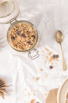 Crunchy granola with dark chocolate chunks in a glass jar set on a white fabric background for a healthy and nutritious morning breakfast meal