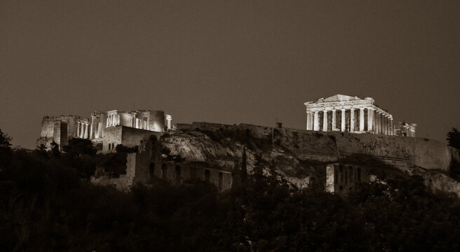 Black and white night view of the Acropolis and Parthenon in Athens, Greece, dramatically illuminated above the city and ancient ruins.
