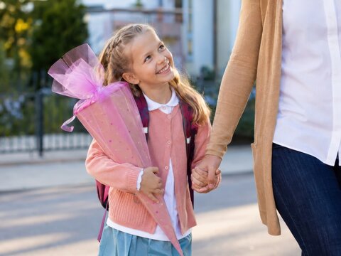 Ein kleines M&auml;dchen h&auml;lt beim Schulweg zur Einschulung in Deutschland die Hand eines Elternteils und blickt voller Vertrauen und Freude auf ihren ersten Schultag.