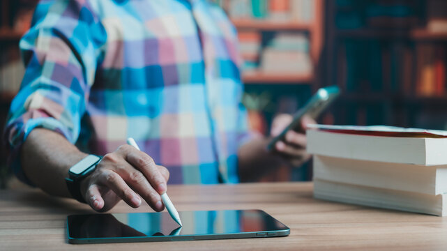 Male student wearing colorful plaid shirt touches digital tablet screen holding stylus pen near stacked books gripping smartphone inside cozy library room, studying online course material sitting.