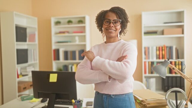 Young african american woman stands with arms crossed by computer monitor and desk piled with files and lamp in an office building; confidence poise.