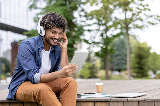 Young man with curly hair smiling while wearing white headphones and watching content on a digital tablet, sitting on wooden steps outdoors with a coffee cup and books nearby