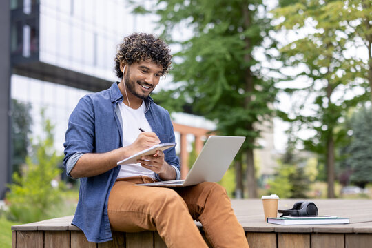 Young man with curly hair engaging in e-learning and remote work outdoors, happily writing in a notebook while listening with earphones and using a laptop