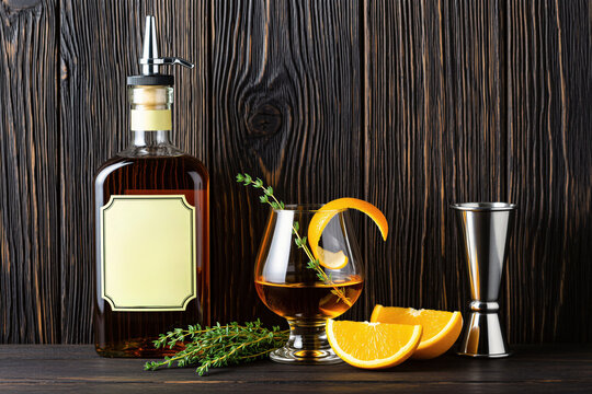 Bottle of amber spirit with glass garnish orange thyme. Bar tools jigger herbs sit on dark wood table. Beverage served with citrus garnish and fresh herbs for enjoyment.