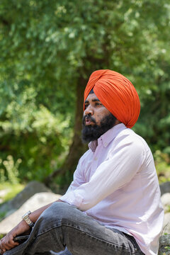 Vertical side view of a young Sikh man wearing an orange turban sitting with a shocked and surprised expression against a blurred background of lush green trees in Himachal Pradesh.