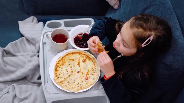 Unhealthy food for children. A girl eats pancakes with gusto while sitting on couch at home and watching TV. Unhealthy food for children. Flour pancakes and berry jam. Nutrition for preschool children