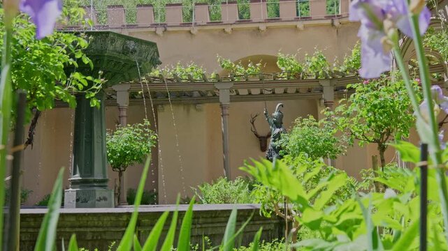 Scenic Historical Castle Garden Patio with actively flowing cast-iron fountain, ornate bronze statue, mounted deer head, and vine-covered arbor on a sunny day in Germany.