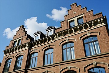 Obraz premium Classic red brick building with gables, large windows stands against clear blue sky. Traditional urban architecture features historical masonry detail with decorative roof structure. Old city facade