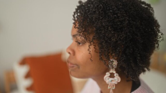 Young woman smiles with tilted head, showing dangling earring while seated on bed in building interior; gentle contentment.
