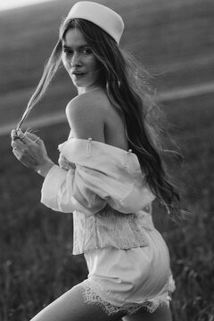 Young woman posing outdoors in a field, wearing a pillbox hat and delicate lace outfit, with long hair blowing in the wind