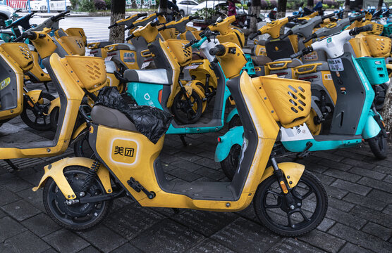 Chongqing, China - 8 Jun 2026 - A row of colorful shared electric scooters and mopeds, including yellow Meituan and turquoise Didi Qingju models, parked neatly on a brick sidewalk along an urban stree