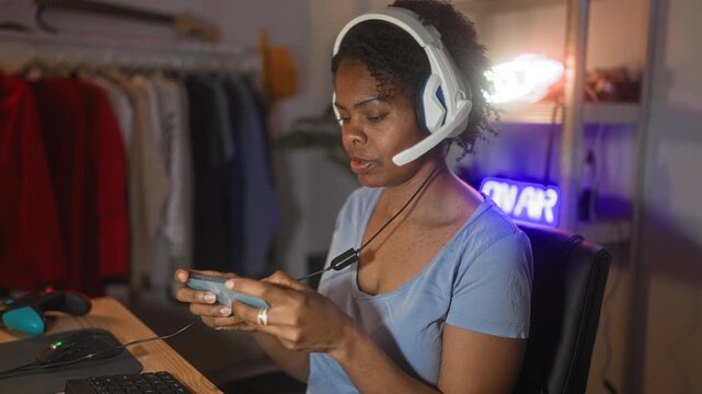 Young woman wearing white headset holds a smartphone like a handheld controller, thumbs tapping controls while seated at a desk with keyboard and mouse in a studio; gaming streaming focus.