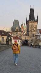 Obraz premium A traveler walks leisurely over Charles Bridge in Prague, surrounded by captivating historical buildings and charming views. The vibrant evening light enhances the beauty of this iconic city.