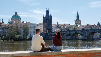 Obraz premium A couple sits peacefully by the Vltava River, admiring beautiful views of Prague. The iconic Charles Bridge and charming buildings create a perfect backdrop for a romantic holiday.