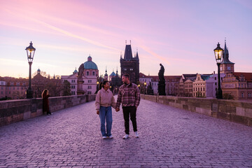 Obraz premium A couple walks hand in hand across the iconic Charles Bridge in Prague during a stunning sunset. The vibrant colors reflect the romantic essence of the city, perfect for tourists exploring its charm.