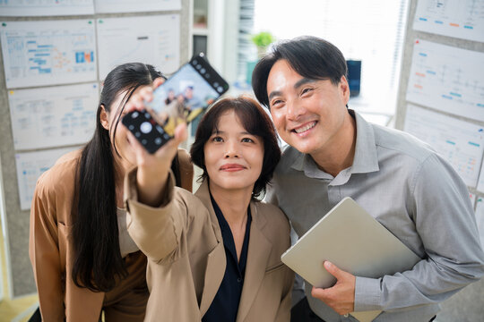 Joyful Asian colleagues smiling and taking a group selfie with a smartphone, enjoying a fun break time together in their modern workspace.