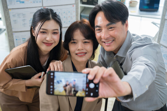 Three happy Asian business people looking at a smartphone camera to take a selfie, capturing a friendly teamwork moment in the office.