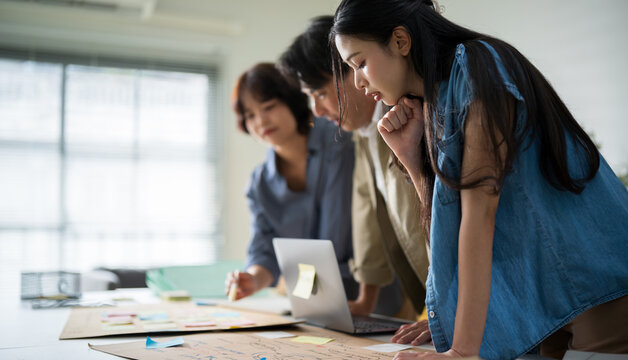 Professional Asian business team gathering around desk to analyze data on laptop screen.