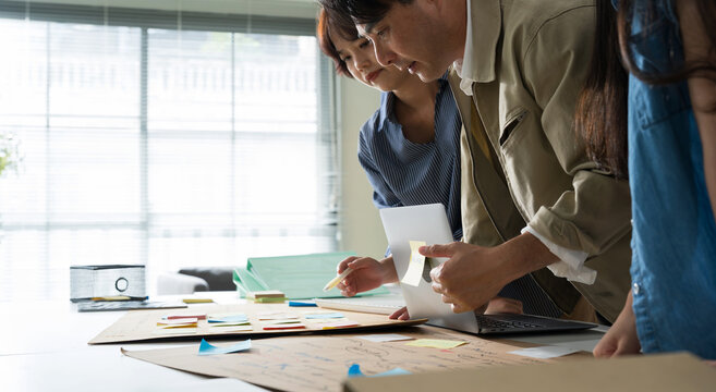 Asian colleagues leaning over a desk, actively brainstorming and sharing creative ideas using colorful sticky notes for project planning.