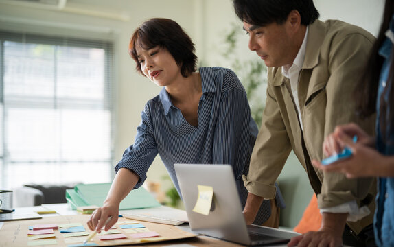 Three Asian corporate workers discussing business strategy, pointing at a laptop screen to share information and solve problems together.