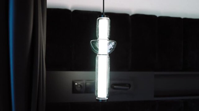 Close up view of a modern vertical glass pendant light fixture glowing brightly in a dark minimalist room. The shot highlights contemporary interior lighting design, elegant home decor, and cozy
