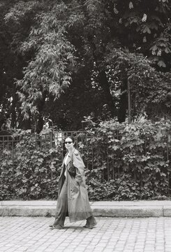 Woman in trench coat walking beside lush urban greenery