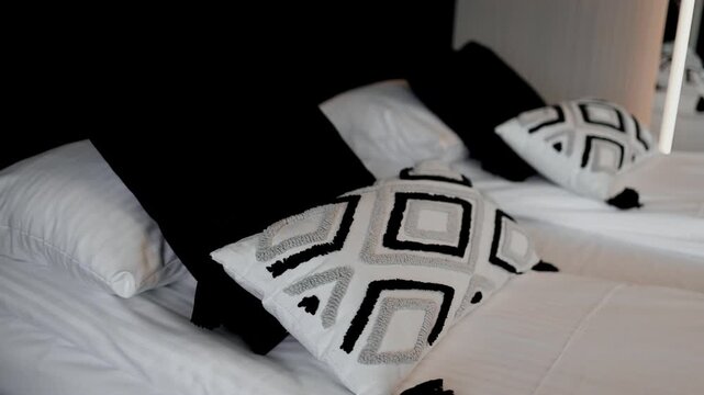 Close up view of a neatly made bed featuring soft white sheets, pillows, and decorative throw pillows with a black and white geometric square pattern. The scene captures modern bedroom interior design