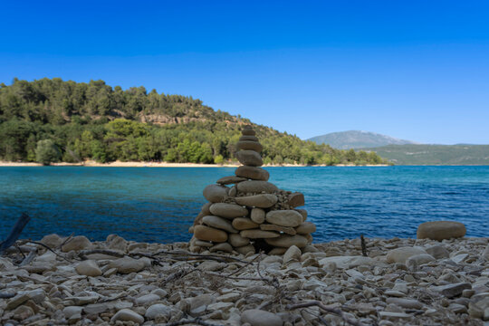 Sainte-Croix-du-Verdon, France - 07 05 2026: Detail view of the Lake Sainte-Croix, Cairn made of stones and pebbles, beach, hills and forests in sunny blue sky