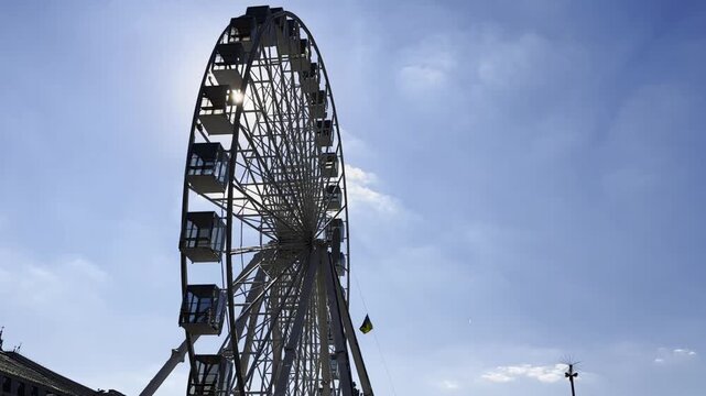 View of a ferris wheel against a clear blue sky with sunlight at background. The sun rays pass through the structure, creating bright highlights and a vibrant summer atmosphere around the attraction