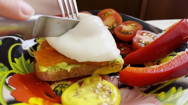 Avocado and red fish sandwich topped with poached egg on home table. Female arms use knife and fork to cut into egg and golden yolk flows over toast highlights appetizing detail of homemade breakfast